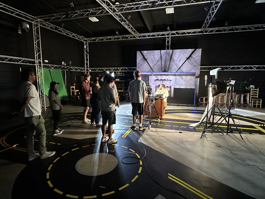 College students work on a set for short film powered by virtual production technology. A large LED screen is in the background, showing the interior of a house.