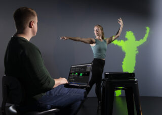 A college student dances in front of a screen that shows her silouette in a green visual pattern. A student in the foreground is working on a computer that is connected to the visual effects.