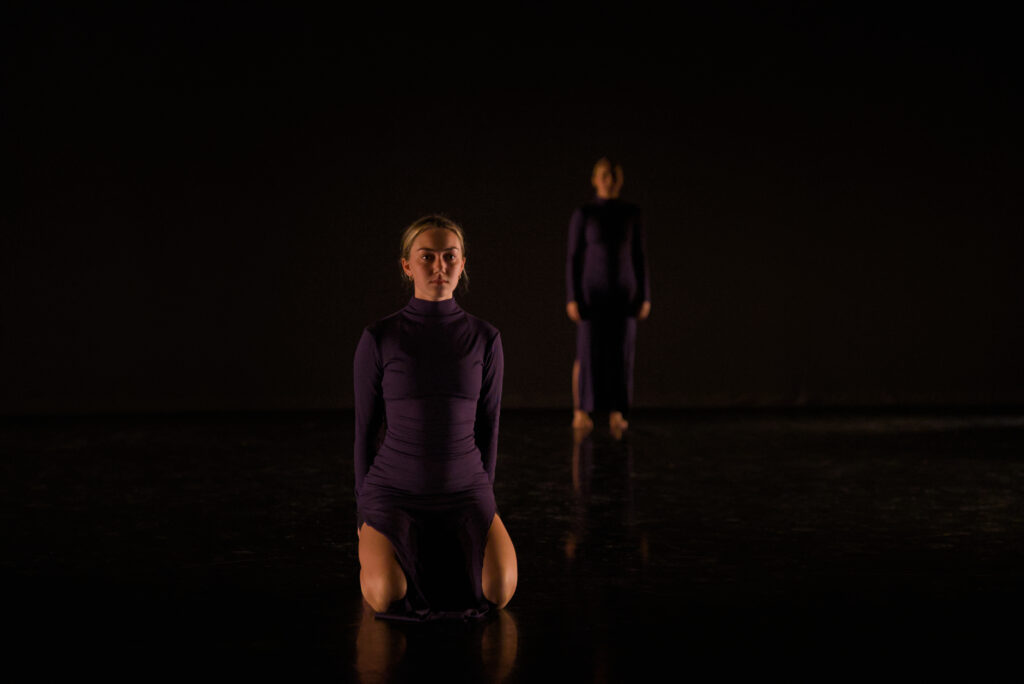 Two college dance students perform in a darkened black box theater, in front of a black background. Both are dressed in black, with one standing in the background and one kneeling in the foreground.