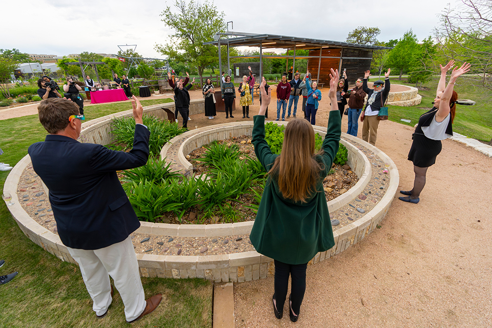 A group of people, some wearing 3D glasses, stand in a circle in an outdoor setting and raise both arms.