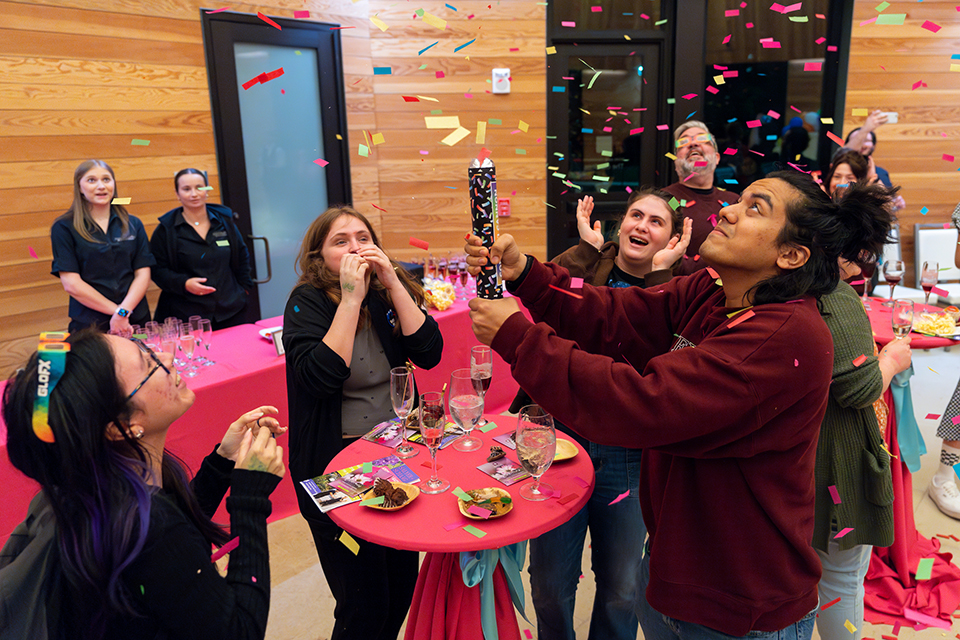 A crowd of people smile and look up as a student pops confetti.