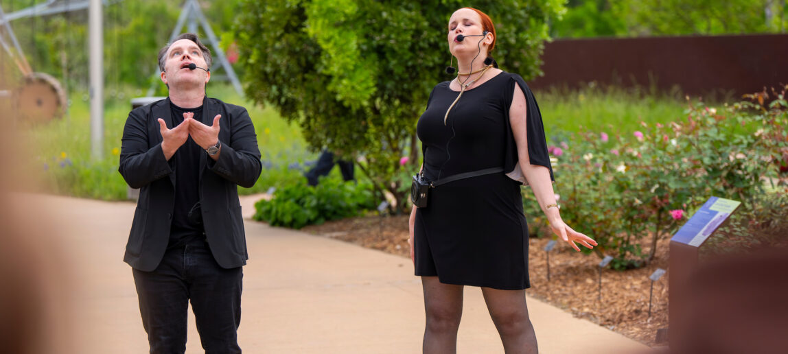 Two artists wearing all black and with headset microphones perform in an outdoor setting.