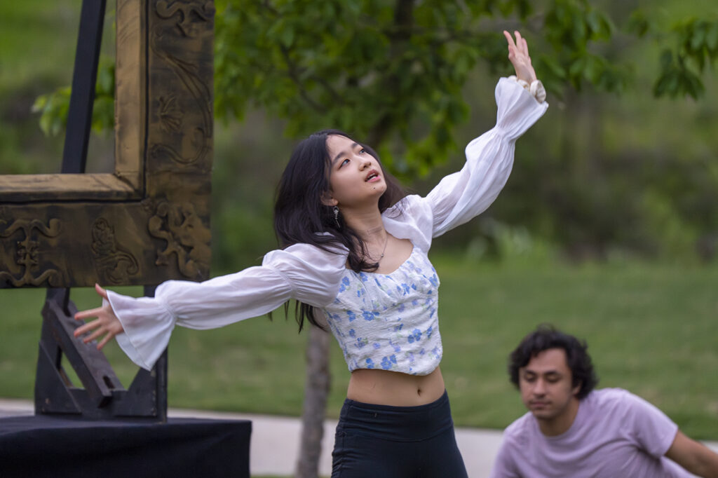 A student stretches out both arms and looks up as she performs in an outdoor setting.
