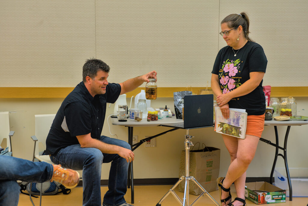 A man holds a glass jar filled with herbal tea, while a woman to his left smiles.