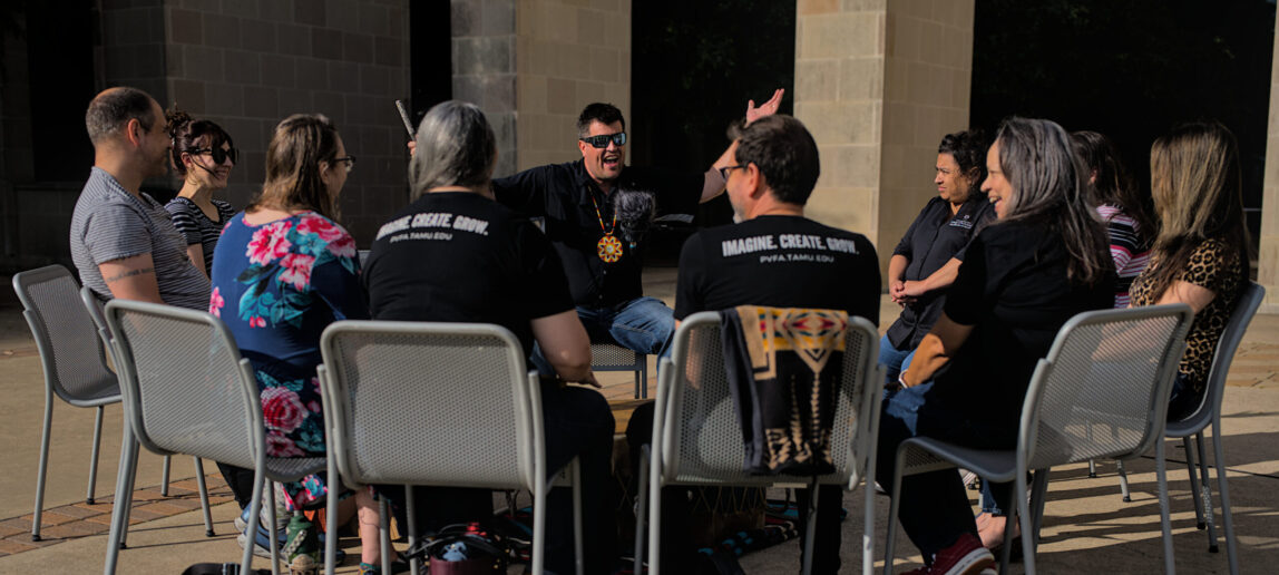 A group of people sit in an outdoor setting. In the middle is a man wearing sunglasses, raising his arms and smiling.