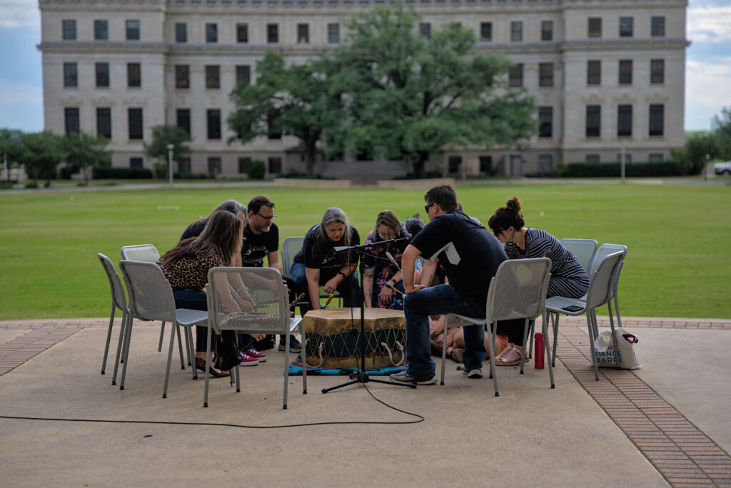 A group of people sit in an outdoor setting, crouched over and playing a ceremonial drum.